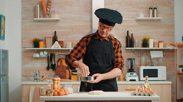 padeiro com bonete e avental usando metálico peneira preparando padaria produtos às lar. feliz idosos chefe de cozinha com cozinha uniforme misturando, aspersão, peneiramento cru ingredientes para cozimento tradicional pão foto