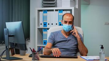 trabalhador autonomo com proteção mascarar falando em Móvel telefone com parceiros, sentado em escrivaninha dentro escritório quarto durante pandemia. trabalhador autonomo trabalhando dentro Novo normal escritório conversando escrevendo Falando em Smartphone foto