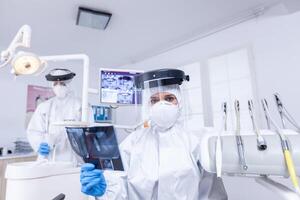 paciente pov dentro dental escritório do dentista discutindo tratamento do dentes cavidade. estomatologia especialista vestindo protetora hazmat terno contra coroavírus mostrando radiografia. foto