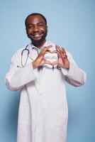 sorridente cardiologista faz uma coração forma com dele dedos para demonstrar preocupação para cuidados de saúde e pacientes. feliz médico médico dentro laboratório casaco com estetoscópio fazer a amor mão gesto. foto