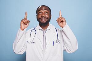 retrato do africano americano médico dentro branco laboratório casaco e estetoscópio, apontando acima com índice dedos dentro estúdio. sorridente médico praticante gesticulando para cima dentro frente do azul fundo. foto