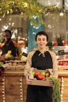 jovem feliz mulher fruta e vegetal mercado impedir proprietário segurando fresco orgânico produzir caixa dentro mãos e sorridente às Câmera. fêmea agricultor vendendo saudável local Comida a partir de beira da estrada ficar de pé foto