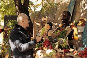 orgânico Comida para idoso. feliz Senior homem falando com fornecedor enquanto compras às local agricultores mercado, jovem amigáveis africano cara agricultor em pé atrás fruta e vegetal impedir e servindo cliente foto