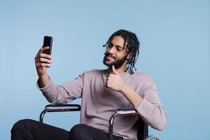 jovem árabe homem falando dentro vídeo ligar em Smartphone e mostrando polegar acima enquanto sentado dentro cadeira de rodas. pessoa com fisica incapacidade tendo conectados comunicação em Móvel telefone foto