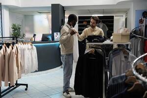 roupas loja cliente segurando camisa para corpo e Perguntando moda consultor para conselho. africano americano homem tentando em formal vestuário enquanto compras e obtendo Socorro a partir de boutique trabalhador foto