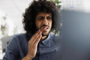 jovem árabe empregado com dor de dente tendo conectados bate-papo com dentista às ambiente de trabalho. empreendedor com dente dor chamando médico em computador portátil enquanto trabalhando dentro o negócio companhia escritório foto