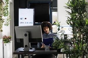 árabe homem analisando relatório em prancheta e trabalhando em computador dentro o negócio aberto espaço. jovem financeiro analista verificação companhia projeto dados às moderno verde escritório local de trabalho foto