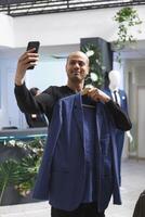 alegre árabe homem levando selfie e exibindo equipamento usando Smartphone frente Câmera enquanto compras dentro boutique. blogueiro segurando Jaqueta e viver transmissão em Móvel telefone dentro roupas loja foto