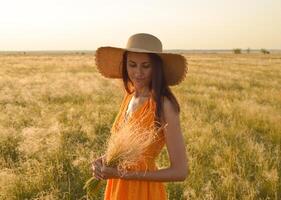 jovem mulher dentro a laranja vestir e uma Palha chapéu em pé em uma campo dentro a raios do a configuração Sol foto