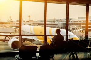 Bangkok cidade, tailândia, 2023 - turista homem estão lendo livros para esperando para a ar avião ao lado a aeroporto terminal janelas com Sol flare e uma amarelo airbus lado de fora fundo. foto