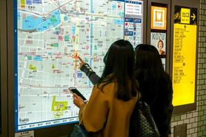 Osaka cidade, Japão, 2023 - chinês mulher turistas olhando às e encontrar atrações do Osaka cidade dentro uma de osaka metrô cidade mapa borda às numba metrô estação. foto