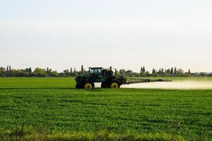 trator com a Socorro do uma pulverizador sprays líquido fertilizantes em jovem trigo dentro a campo. foto