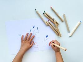 pequeno criança desenha com colori lápis em papel em branco mesa. foto