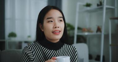 retrato do relaxante jovem mulher segurando copo apreciar cheiro café ou chá e bebendo com felicidade dentro manhã enquanto sentar em sofá dentro vivo quarto, grátis tempo, leve pausa dentro casa, sorrindo foto