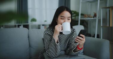 retrato do feliz jovem ásia mulher sentar em sofá apreciar conversando em Móvel telefone enquanto bebendo café dentro manhã dentro vivo quarto, grátis tempo, leve quebrar, sorrindo foto