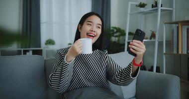 retrato do feliz jovem ásia mulher sentar em sofá segurando café copo e apreciar levando selfies em Móvel telefone às casa livre tempo, leve quebrar, sorrindo foto