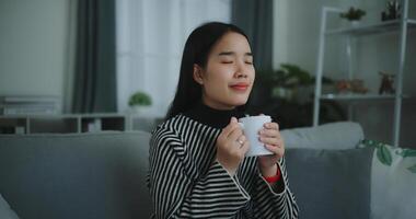 retrato do relaxante jovem mulher segurando copo apreciar cheiro café ou chá e bebendo com felicidade dentro manhã enquanto sentar em sofá dentro vivo quarto, grátis tempo, leve pausa dentro casa, sorrindo foto