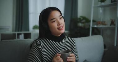 retrato do feliz jovem ásia mulher apreciar ouvindo para a música com sem fio fones de ouvido enquanto sentado beber café em sofá dentro vivo quarto às lar, lazer e estilo de vida, grátis Tempo foto