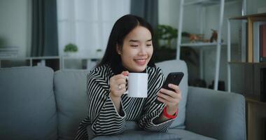 retrato do feliz jovem ásia mulher sentar em sofá apreciar conversando em Móvel telefone enquanto bebendo café dentro manhã dentro vivo quarto, grátis tempo, leve quebrar, sorrindo foto