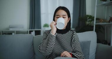 retrato do relaxante jovem mulher segurando copo apreciar cheiro café ou chá e bebendo com felicidade dentro manhã enquanto sentar em sofá dentro vivo quarto, grátis tempo, leve pausa dentro casa, sorrindo foto