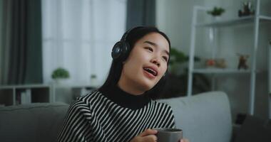 retrato do feliz jovem ásia mulher apreciar ouvindo para a música com sem fio fones de ouvido enquanto sentado beber café em sofá dentro vivo quarto às lar, lazer e estilo de vida, grátis Tempo foto