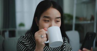 retrato do feliz jovem ásia mulher sentar em sofá apreciar conversando em Móvel telefone enquanto bebendo café dentro manhã dentro vivo quarto, grátis tempo, leve quebrar, sorrindo foto