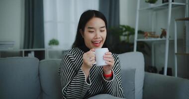 retrato do relaxante jovem mulher segurando copo apreciar cheiro café ou chá e bebendo com felicidade dentro manhã enquanto sentar em sofá dentro vivo quarto, grátis tempo, leve pausa dentro casa, sorrindo foto