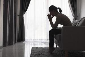 silhueta do uma pessoa sofrimento a partir de depressão dentro a casa, depressivo mulher sentado sozinho em a sofá sentir estresse, triste e preocupado dentro a Sombrio sala. pessoa estão estressado. foto