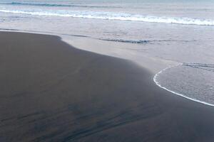 oceano de praia com Preto vulcânico areia, a céu é refletido dentro a enrolado costas onda do a surfar foto