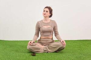 jovem mulher sentado dentro meditação pose e parece acima sonhadoramente em luz fundo foto