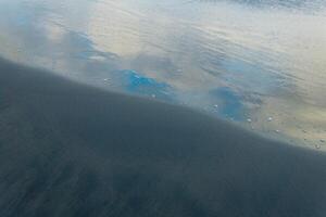 oceano de praia com Preto vulcânico areia, a céu é refletido dentro a enrolado costas onda do a surfar foto