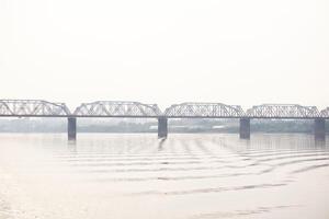 Ferrovia ponte sobre uma Largo rio e a silhueta do uma distante cidade atrás isto dentro a manhã névoa foto