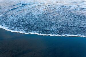 oceano de praia com Preto vulcânico areia, a céu é refletido dentro a enrolado costas onda do a surfar foto