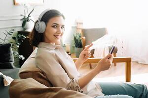 alegre jovem mulher com sem fio fones de ouvido sorrisos, pontos para Smartphone, lendo uma mensagem. positivo e feliz fêmea desfrutando tecnologia e comunicação com sem fio fones de ouvido. foto