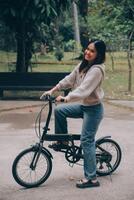feliz jovem ásia mulher enquanto equitação uma bicicleta dentro uma cidade parque. ela sorriu usando a bicicleta do transporte. ambientalmente amigáveis conceito. foto