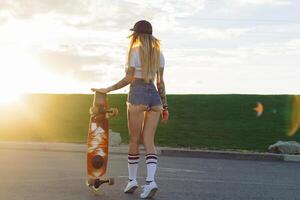 retrato do uma hipster jovem menina sorridente com uma Longboard às pôr do sol. foto