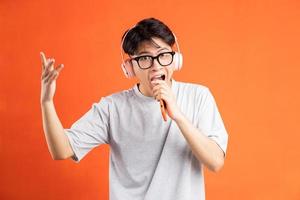 retrato de jovem asiático segurando um telefone inteligente na mão, usando fones de ouvido e cantando, isolado em um fundo laranja foto