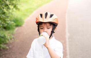 saudável criança Garoto vestindo uma bicicleta capacete e bebendo água, criança com sede Garoto segurando uma garrafa do água vestindo uma ciclismo capacete equitação uma bicicleta dentro a parque, conceito para segurança, infância saúde Cuidado foto