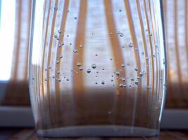 uma curvado transparente vidro preenchidas com levemente carbonatado mineral água em a fundo do uma rattan cortina em peitoril da janela foto