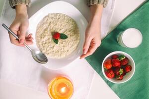 aveia com leite. mingau com leite. cereais, delicioso café da manhã, dieta café da manhã. vela. morangos, aveia e vidro do leite. mãos do uma jovem mulher com uma colher. saudável comendo cocept foto