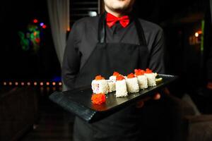 homem dentro Preto camisa e vermelho arco gravata segurando bandeja do Sushi foto