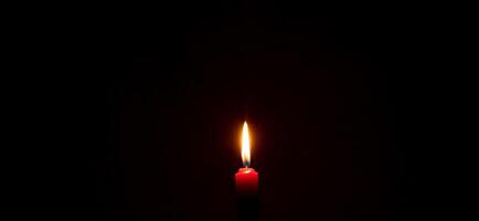 uma solteiro queimando vela chama ou luz brilhando em uma vermelho vela em Preto ou Sombrio fundo em mesa dentro Igreja para Natal, velório ou memorial serviço com cópia de espaço foto