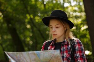 jovem caucasiano mulher dentro uma chapéu olhando às uma geográfico mapa dentro a sombra do árvores dentro uma parque ou floresta. Alto qualidade foto