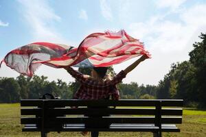 jovem menina senta em Banco e detém a americano bandeira acenando dentro a vento. Alto qualidade foto