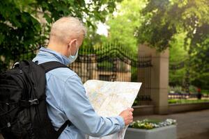 a grisalho homem dentro uma jeans camisa e com uma mochila parece às a mapa. mascarar em a face. cidade rua. cerca e parque estão visível. uma Novo viagem conceito dentro a Novo realidades dentro público lugares. Alto foto