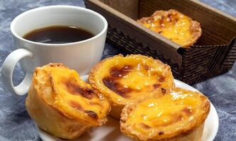 grande quantidade do sobremesas pastel de nata ou Português ovo azedo com uma branco copo do Preto café. pastel de belm é uma pequeno torta com uma crocantes sopro pastelaria crosta e uma creme creme enchimento. foto