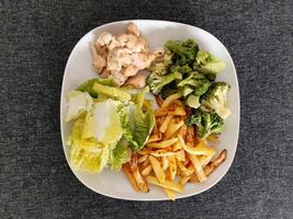 caseiro grelhado frango com francês fritas, brócolis e verde salada, servido em uma branco prato foto