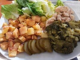 caseiro grelhado frango com francês fritas, verde salada, brócolis e pepino picles servido em uma branco prato foto