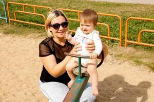 mãe rolos bebê filha em uma balanço em a Parque infantil foto