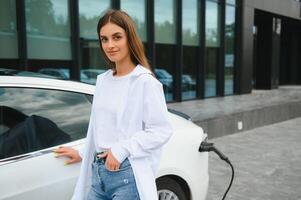 feliz jovem adulto mulher sorridente largo, olhando ausente, cobrando automóvel bateria a partir de pequeno público estação, em pé perto elétrico carro. foto